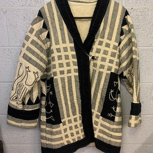 May include: A black and white quilted jacket with a geometric pattern and embroidered birds. The jacket has a black collar and a single button closure.