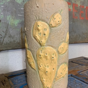 May include: A cylindrical lamp with a textured, light gray surface. The lamp features raised, yellow-green cactus designs. The lamp has a beige neck and a green cord. The lamp is on a wooden surface.