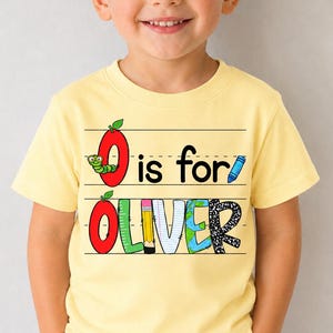 May include: A pale yellow short-sleeve t-shirt featuring a graphic design with the text O is for OLIVER. The design includes a red apple with a green caterpillar, a blue crayon, and the name OLIVER in colorful, patterned block letters.