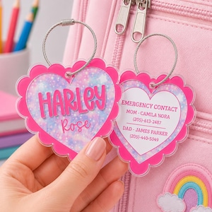 May include: Two heart-shaped bag tags with bright pink scalloped edges and silver wire loops. One tag features the name Harley Rose in pink text over a glittery pastel background. The other tag displays emergency contact information in pink text.