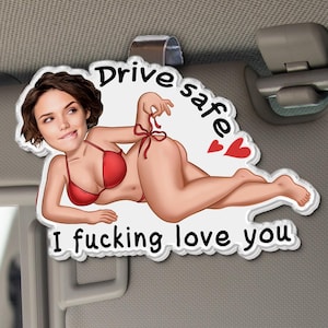 May include: A white car visor accessory with a cartoon illustration of a woman in a red bikini. The text on the accessory reads "Drive safe" and "I fucking love you". Two red hearts are also included in the design.