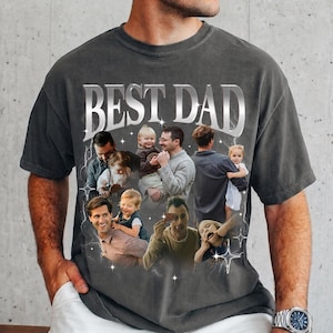 May include: Dark gray t-shirt with the text "BEST DAD" in a silver, metallic font. The shirt features a collage of photos of a father with his children. The photos are surrounded by lightning bolt and star graphics.