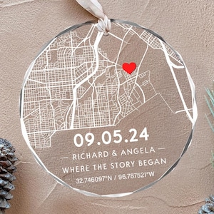 May include: Clear acrylic ornament with a map design, a red heart, and the text "09.05.24 RICHARD & ANGELA - WHERE THE STORY BEGAN 32.746097°N / 96.787521°W". The ornament is round and has a ribbon for hanging.