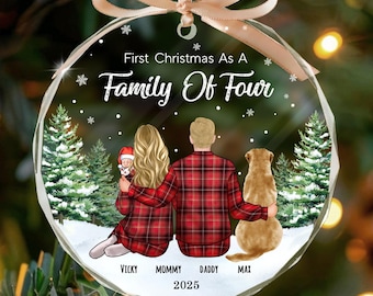 May include: A clear, faceted Christmas ornament with a family portrait. The ornament features a couple, a baby, and a dog, with the text "First Christmas As A Family Of Four" and names. A beige ribbon is attached for hanging.