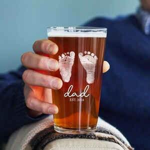 May include: A clear glass pint glass with a brown beer inside. The glass is etched with two baby footprints and the text "dad est. 2024".