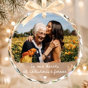 May include: A circular glass Christmas ornament featuring a photo of an older person and a younger person embracing in a field of yellow flowers. The ornament has a faceted edge, a cream-colored ribbon bow, and the text In our hearts at Christmas & forever.