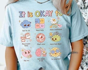Puede incluir: Una camiseta azul claro con el texto It is OKAY to y nueve ilustraciones coloridas con los pies de foto: Not be ok, Need help, Make mistakes, Need more time, Start over, Have bad days, Say no, Not know it all y Be yourself.