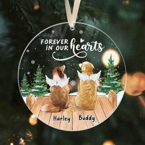 May include: A clear, round Christmas ornament with the text "FOREVER IN OUR hearts". It features illustrations of two dogs with angel wings, named Harley and Buddy, on a wooden dock with a snowy background and Christmas trees.