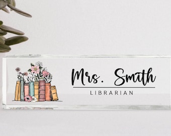 Puede incluir: Una placa de nombre de escritorio de vidrio transparente con un diseño floral y una pila de libros. El texto "Mrs. Smith LIBRARIAN" está impreso en la placa.