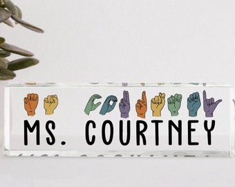 May include: A clear acrylic desk nameplate with the text "MS. COURTNEY" in black lettering. The name is spelled out in American Sign Language with colorful hand gestures.