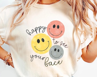 May include: A cream-colored t-shirt featuring a graphic of three smiley faces in coral, yellow, and gray, each with a subtle floral pattern. The text "happy to see your face" is printed in black cursive script around the smiley faces.