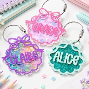 May include: Three acrylic luggage tags with metal cable loops, each shaped like a scalloped shell with a bow at the top. The tags feature glittery backgrounds and raised lettering reading GRACE in pink, CLAIRE in purple, and ALICE in teal.