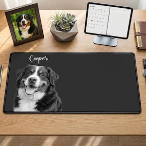 May include: Black desk mat featuring a smiling Bernese Mountain Dog portrait with the name "Cooper" in white script. A framed photo of a similar dog, a succulent plant, and a tablet with a calendar are also on the wooden desk.