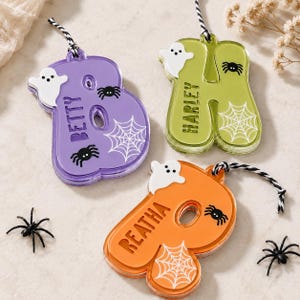 May include: Three acrylic letter ornaments in purple, lime green, and orange, each featuring a white ghost, black spiders, and a white spiderweb graphic. The letters are personalized with the names Betty, Harley, and Reatha, and include black and white striped hanging cords.