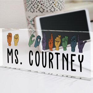 May include: A clear acrylic desk nameplate with the text "MS. COURTNEY" in black lettering. Above the name, there are rainbow-colored hand signs spelling out the name "COURTNEY" in American Sign Language.
