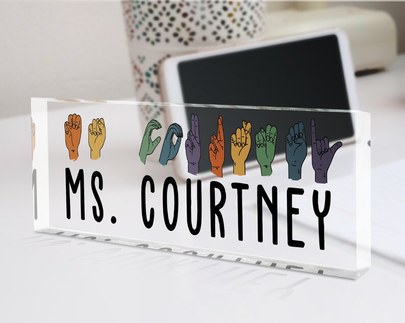 May include: A clear acrylic desk nameplate with the text "MS. COURTNEY" in black lettering. Above the name, there are rainbow-colored hand signs spelling out the name "COURTNEY" in American Sign Language.