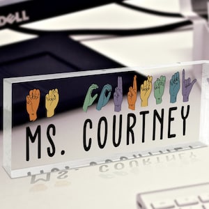 May include: A clear acrylic desk nameplate with the text "MS. COURTNEY" in black lettering. The nameplate features a row of colorful hand signs representing the letters of the name.