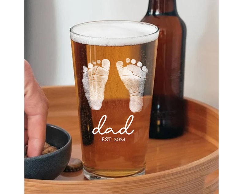 May include: A clear glass pint glass with a design of two baby footprints and the text "dad est. 2024".