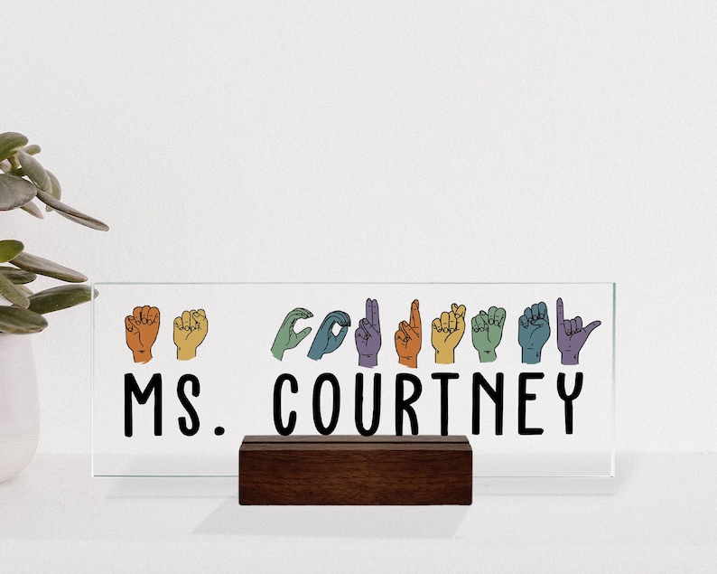 May include: A glass desk sign with a wooden stand. The sign features the name "MS. COURTNEY" in black text and a row of colorful hands signing the letters of the name in American Sign Language.