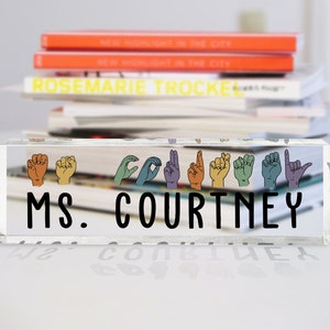 May include: A clear acrylic desk sign with the text "MS. COURTNEY" in black lettering. The sign features a row of colorful hands in American Sign Language spelling out the name "COURTNEY".