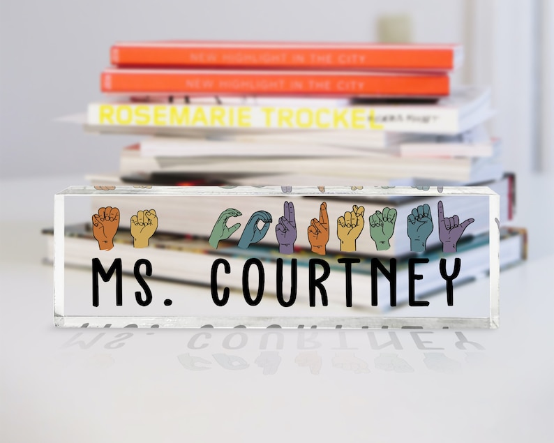May include: A clear acrylic desk sign with the text "MS. COURTNEY" in black lettering. The sign features a row of colorful hands in American Sign Language spelling out the name "COURTNEY".