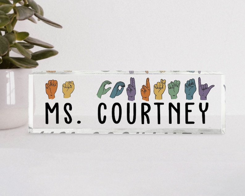 May include: A clear acrylic desk nameplate with the text "MS. COURTNEY" in black lettering. The name is spelled out in American Sign Language with colorful hand gestures.