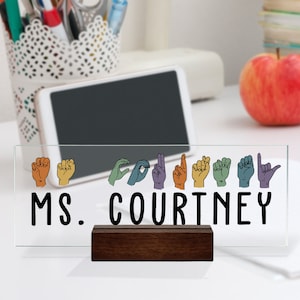 May include: A clear glass desk sign with a wooden stand. The sign features a row of colorful hand signs spelling out "MS. COURTNEY".