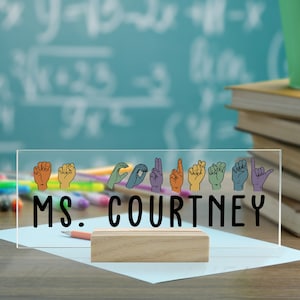 May include: A clear acrylic desk sign with the text "MS. COURTNEY" in black lettering. The sign features a row of colorful hand gestures in American Sign Language.