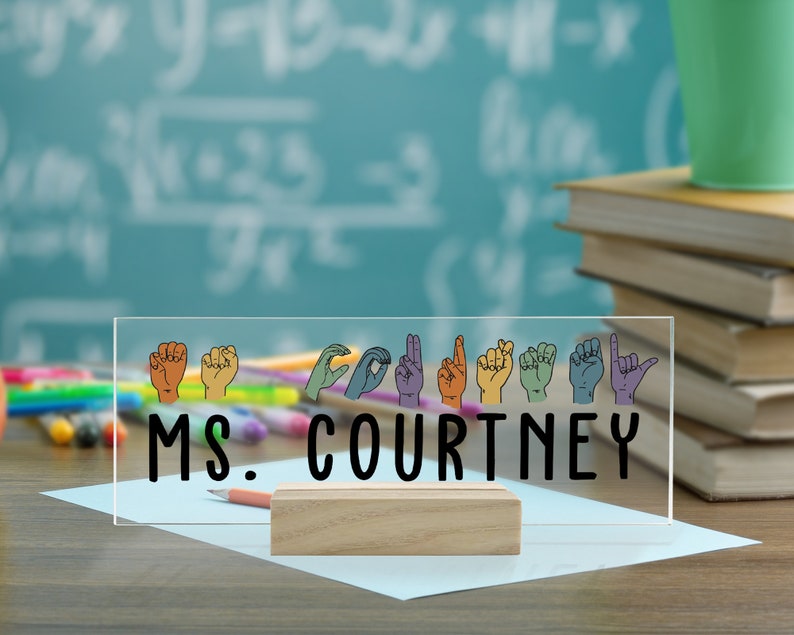 May include: A clear acrylic desk sign with the text "MS. COURTNEY" in black lettering. The sign features a row of colorful hand gestures in American Sign Language.