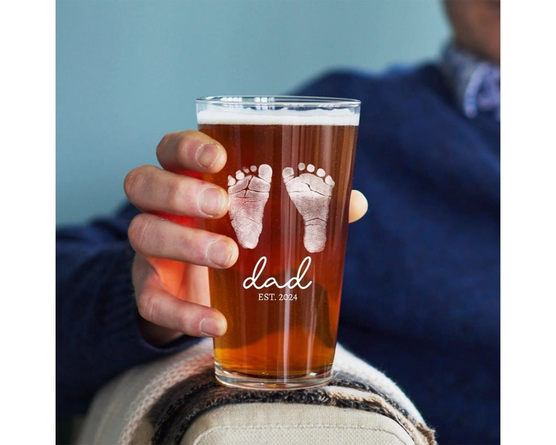 May include: A clear glass pint glass with a brown beer inside. The glass is etched with two baby footprints and the text "dad est. 2024".