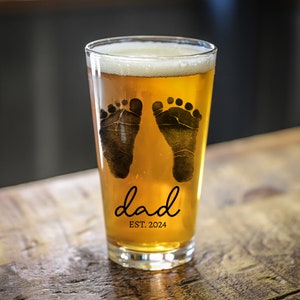 May include: A clear glass pint glass with a design of two black baby footprints and the text "dad est. 2024". The glass is filled with a light amber colored beer.