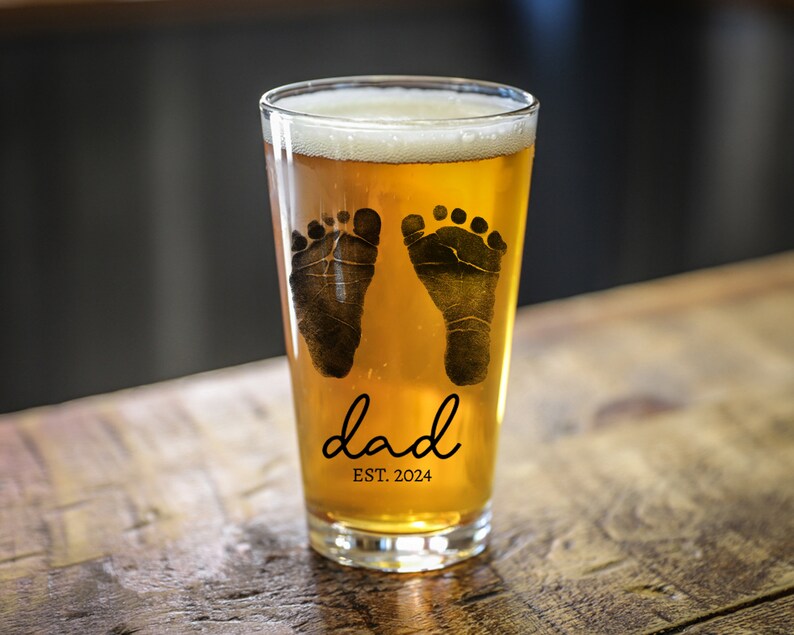 May include: A clear glass pint glass with a design of two black baby footprints and the text "dad est. 2024". The glass is filled with a light amber colored beer.