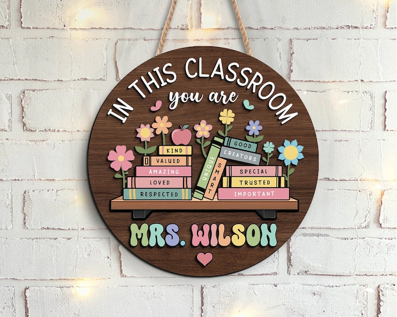 May include: A round wooden sign with the text "In this classroom you are" and a stack of books with positive words like "kind", "valued", "amazing", "loved", "respected", "unique", "smart", "good creators", "special", "trusted", and "important". The sign also has the text "Mrs. Wilson" and a heart.