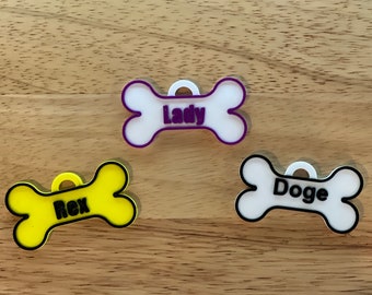 May include: Three bone-shaped dog tags with different names. The top tag is white with a purple outline and says "Lady". The middle tag is yellow with a black outline and says "Rex". The bottom tag is white with a black outline and says "Doge".