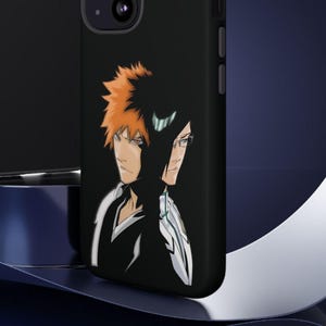 Op de afbeelding: Twee zwarte iPhone-hoesjes met een afbeelding van twee anime-personages. De personages zijn in zwart-wit met oranje haar. Het hoesje aan de linkerkant is een effen zwart hoesje. Het hoesje aan de rechterkant heeft een uitsparing voor de camera.