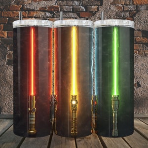 Pode incluir: Três copos de aço inoxidável com designs de sabre de luz de diferentes cores. Os copos estão sobre uma superfície de madeira em frente a uma parede de tijolo.