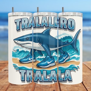 May include: A white tumbler with a blue shark wearing blue sneakers, standing on a beach with waves. The tumbler has the words "TRALALERO TRALALA" in blue. The background shows a blurred ocean scene.