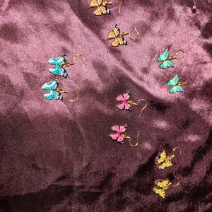 May include: A collection of butterfly earrings in various colors, including yellow, pink, blue, and green. The earrings have gold-colored hooks and are arranged on a dark purple, textured surface. The butterfly wings have black detailing.
