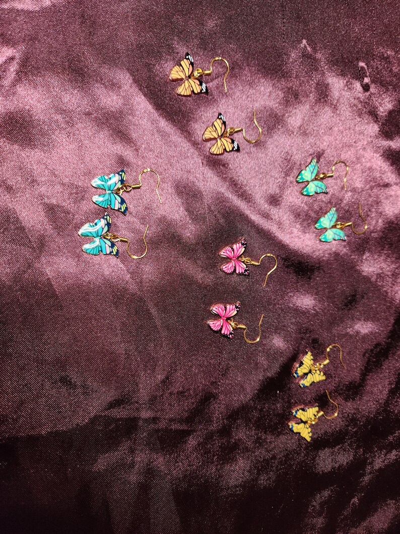 May include: A collection of butterfly earrings in various colors, including yellow, pink, blue, and green. The earrings have gold-colored hooks and are arranged on a dark purple, textured surface. The butterfly wings have black detailing.