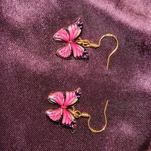 May include: A pair of butterfly earrings. Each earring features a pink butterfly design with black and white accents, set against a gold-colored metal. The earrings have a hook closure.