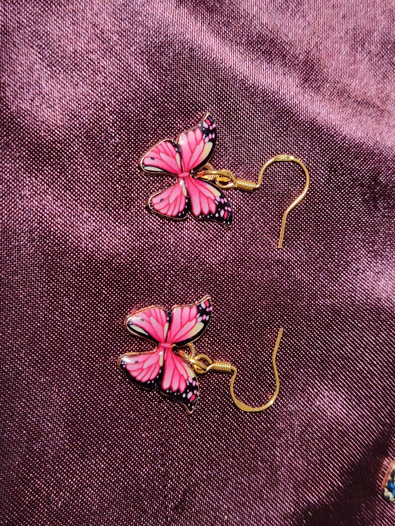 May include: A pair of butterfly earrings. Each earring features a pink butterfly design with black and white accents, set against a gold-colored metal. The earrings have a hook closure.