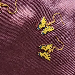 May include: Gold-colored butterfly earrings with yellow wings and blue accents. The butterfly-shaped earrings are hanging on gold-colored hooks. The earrings are set against a dark purple background.