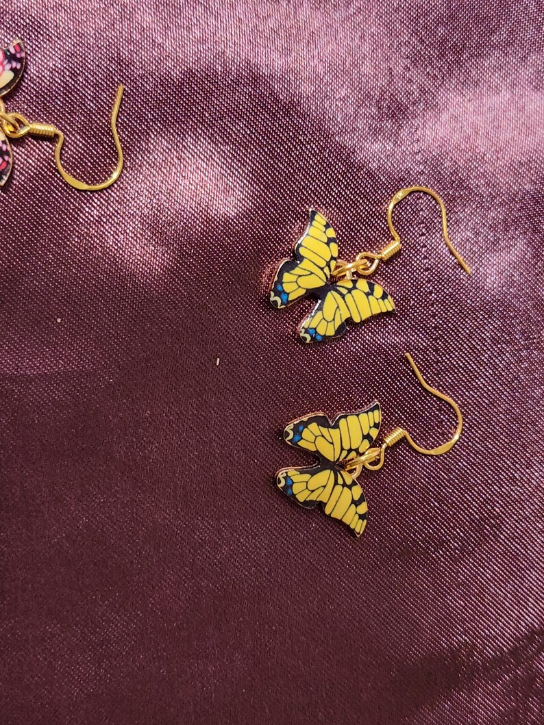 May include: Gold-colored butterfly earrings with yellow wings and blue accents. The butterfly-shaped earrings are hanging on gold-colored hooks. The earrings are set against a dark purple background.