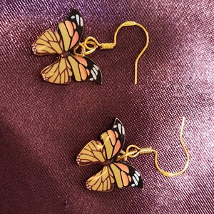 May include: A pair of butterfly earrings. Each earring features a gold-colored fish hook and a butterfly pendant with yellow, orange, black, and white details. The earrings are set against a textured, dark purple background.