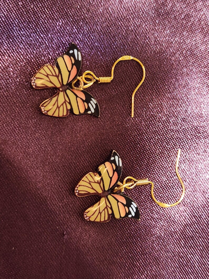 May include: A pair of butterfly earrings. Each earring features a gold-colored fish hook and a butterfly pendant with yellow, orange, black, and white details. The earrings are set against a textured, dark purple background.