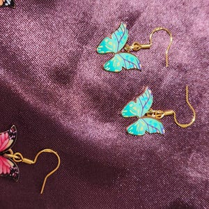 May include: Close-up of butterfly earrings with gold-colored hooks. The butterfly wings are turquoise with purple accents. The earrings are set against a dark purple, textured background.