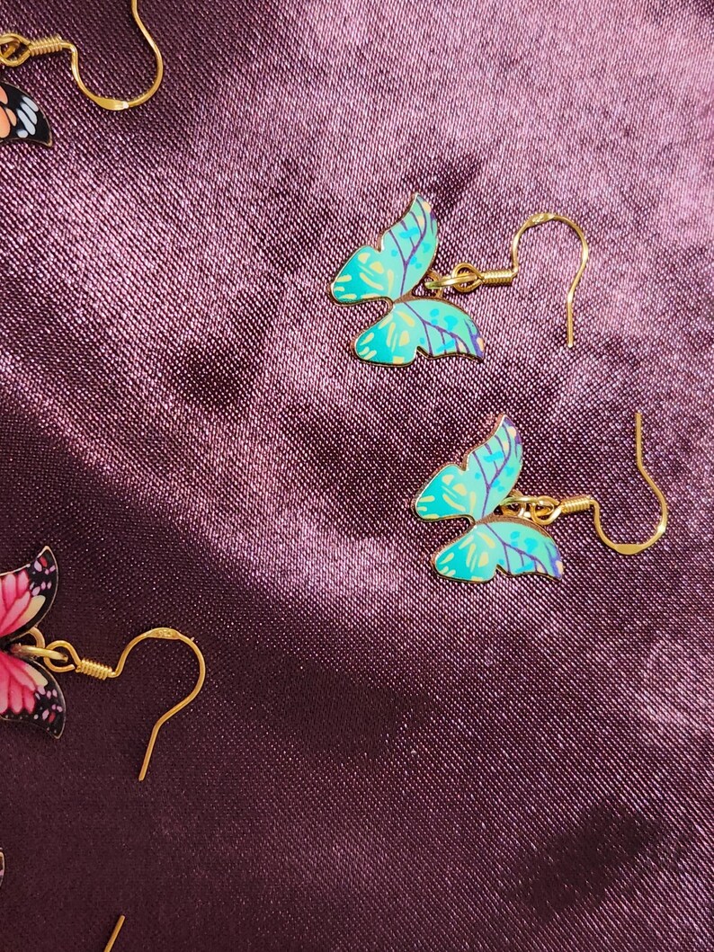 May include: Close-up of butterfly earrings with gold-colored hooks. The butterfly wings are turquoise with purple accents. The earrings are set against a dark purple, textured background.