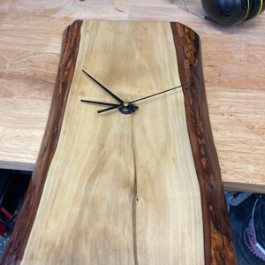 May include: A wooden clock with a natural wood grain finish and black hands. The clock is made from a live edge slab of wood with a dark brown border.