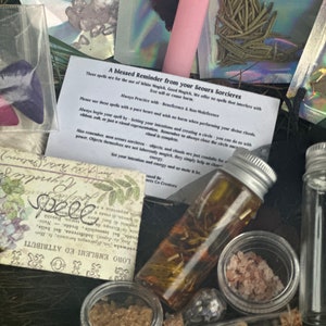 May include: A collection of small glass bottles filled with herbs, crystals, and other ingredients for spell casting. The bottles are arranged on a wooden surface with a handwritten note that reads "A blessed Reminder from your Seours Sorcieres. These spells are for the use of White Magick. Good Magick. We offer no spells that interfere with free will or cause harm. Always Practice with Beneficence & Non-Maleficence."
