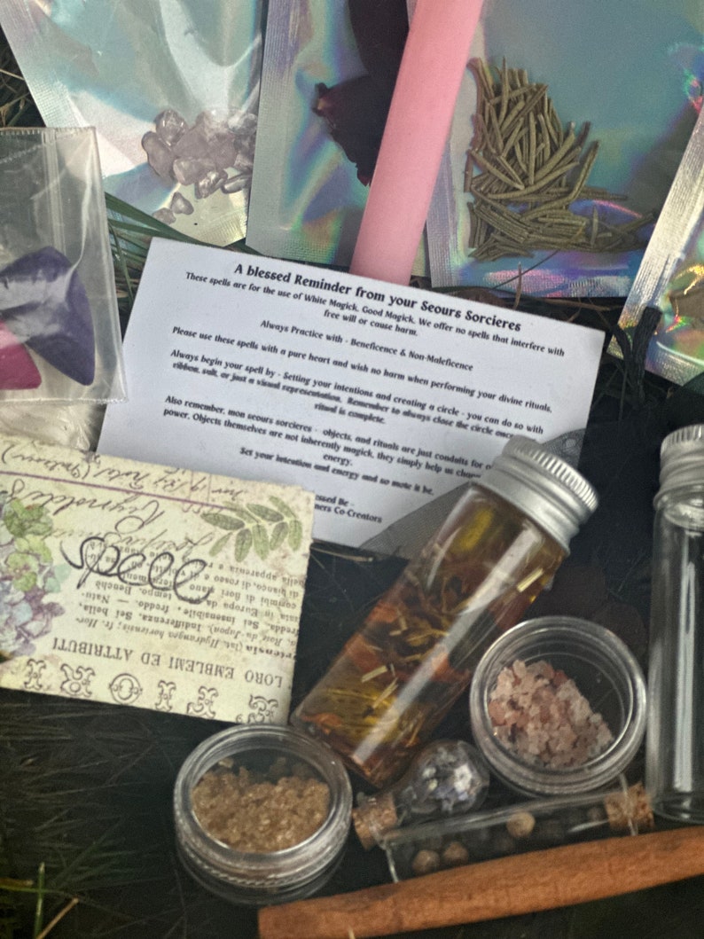 May include: A collection of small glass bottles filled with herbs, crystals, and other ingredients for spell casting. The bottles are arranged on a wooden surface with a handwritten note that reads "A blessed Reminder from your Seours Sorcieres. These spells are for the use of White Magick. Good Magick. We offer no spells that interfere with free will or cause harm. Always Practice with Beneficence & Non-Maleficence."